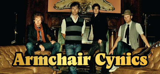 Four men stand and sit in a vintage-style room with brown leather sofas and a fireplace. A large taxidermy animal head is mounted above the mantle. Bold yellow text reads Armchair Cynics.
