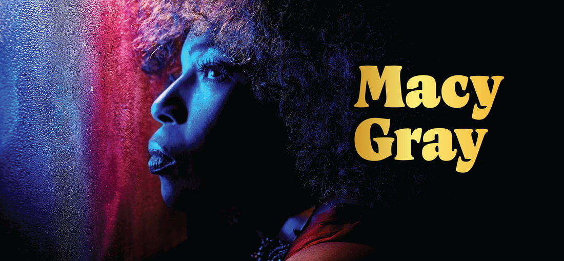 A woman with curly hair is dramatically lit in blue and pink, gazing thoughtfully to the side. The text Macy Gray in bold yellow letters appears beside her on a dark background.