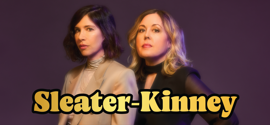 Two women stand against a purple background, one with dark hair in a tan suit and the other with blonde hair in a black top. Large yellow text at the bottom reads Sleater-Kinney.