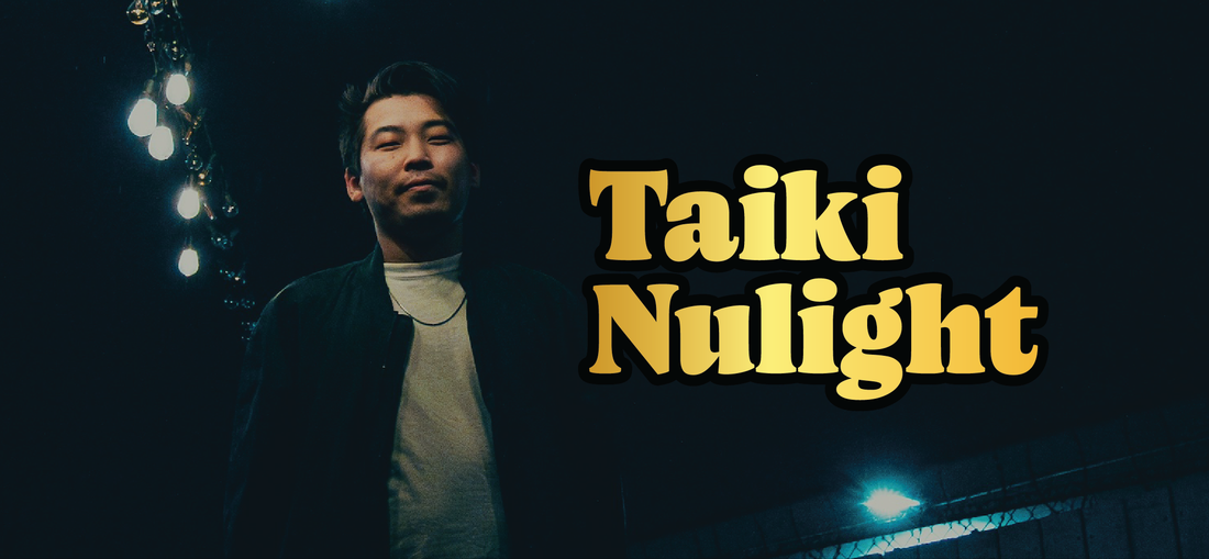 A man stands outdoors at night under bright lights, wearing a dark jacket over a white shirt. Large yellow text next to him reads Taiki Nulight. The background is dark with a few visible lights.