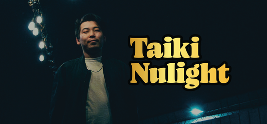 A man stands outdoors at night under bright lights, wearing a dark jacket over a white shirt. Large yellow text next to him reads Taiki Nulight. The background is dark with a few visible lights.