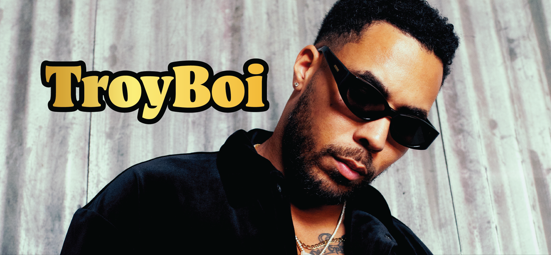 	 A man wearing dark sunglasses and a black jacket stands in front of a wooden background. The name TroyBoi appears in bold yellow and black text on the left side of the image.